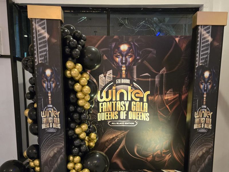 Black and gold chrome balloon garland photo backdrop wall with gold-trimmed columns and a custom printed floor decal, built for an all-black winter fantasy gala in Queens, NY