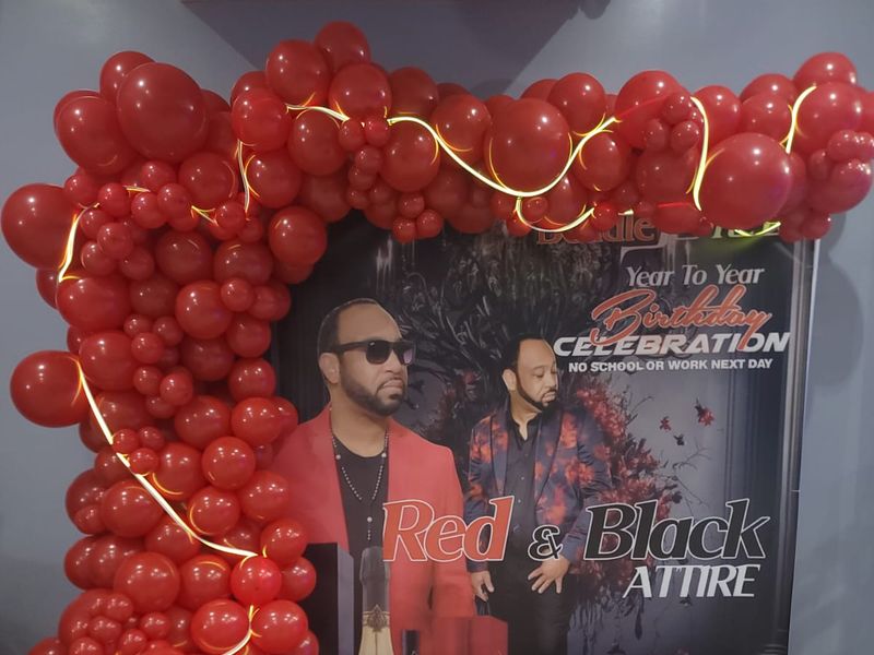 Red balloon garland arch with warm neon rope lighting framing a custom birthday step-and-repeat banner and matching red and black printed floor decal at a Long Island celebration