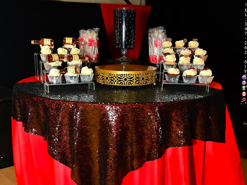 Round dessert table rental dressed in a red satin floor-length linen with a black sequin overlay, holding cupcake towers and a gold cake stand at a Long Island 65th birthday celebration